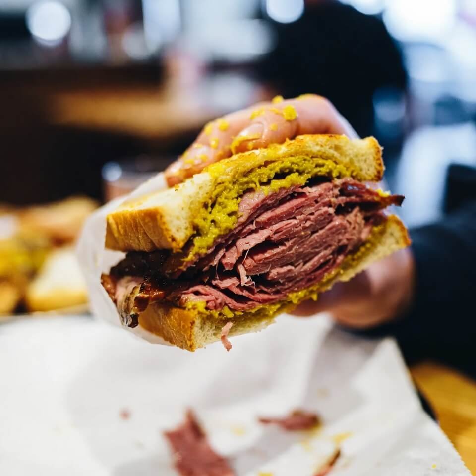 Hand holding messy pastrami sandwich mid-bite, mustard dripping everywhere.